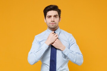 Confident handsome young business man in classic blue shirt posing isolated on yellow background studio portrait. Achievement career wealth business concept. Mock up copy space. Straightening tie.