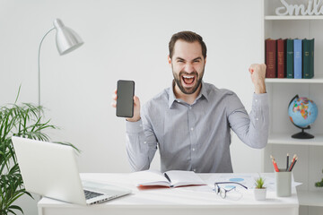 Happy business man in gray shirt sit at desk work on laptop in light office on white wall background. Achievement business career concept. Hold mobile phone with blank screen doing winner gesture.