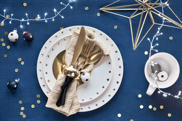 Christmas table setup with white plate and golden utensils and gilded decorations. Flat lay, top view on classic blue. Dark blue linen textile background. Xmas lights garland.