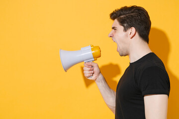Side veiw of angry young man guy in casual black t-shirt posing isolated on yellow background studio portrait. People sincere emotions lifestyle concept. Mock up copy space. Screaming in megaphone.