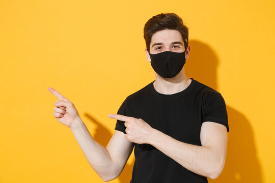 Smiling young man guy in black t-shirt face mask isolated on yellow background studio portrait. Epidemic pandemic coronavirus 2019-ncov sars covid-19 flu virus concept. Pointing index fingers aside.