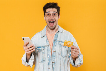 Photo of delighted young man holding credit card and using mobile phone