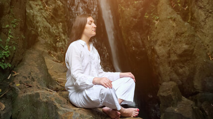 Fototapeta na wymiar concentrated woman with loose dark hair in white costume meditates sitting in yoga lotus position on rock against tropical waterfall closeup