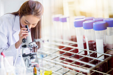 Beautiful Asian lab technician woman assistant analyzing a blood sample in test tube at laboratory with microscope. Medical, pharmaceutical and scientific service, research and development concept.