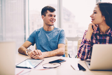 Two successful smiling teenagers casual dressed sitting desktop with netbooks connected to wifi, happy hipster guy looking at cheerful girl at coworking space, concept of technology and communication