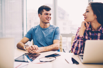 Cheerful male and female colleagues dressed in casual wear laughing during discussion of creative ideas sitting at meeting table in office.Positive employees talking with each other about project