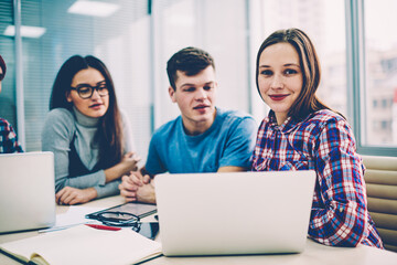 Portrait of cheerful hipster girl looking at camera during studying with friends via computers at college campus, group of positive students using technology and wifi for learning online course.