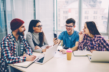Successful smiling hipster guy making video call via smartphone with 4g connection while sitting with friends at coworking space, group of happy students spending time with technology indoors.