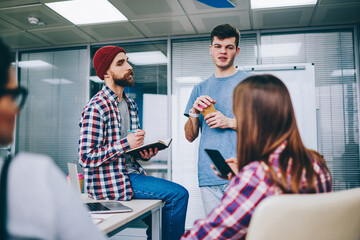 Young handsome hipster guy holding cup of coffee and explaining main ideas of startup project to male and female friends while girl sitting with cellular and chatting during other man notes theses.