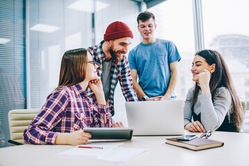 Fototapeta premium Successful group of young professionals in casual wear laughing during cooperation on design project sitting at laptop in office.Positive students teamworking on common task during brainstorming
