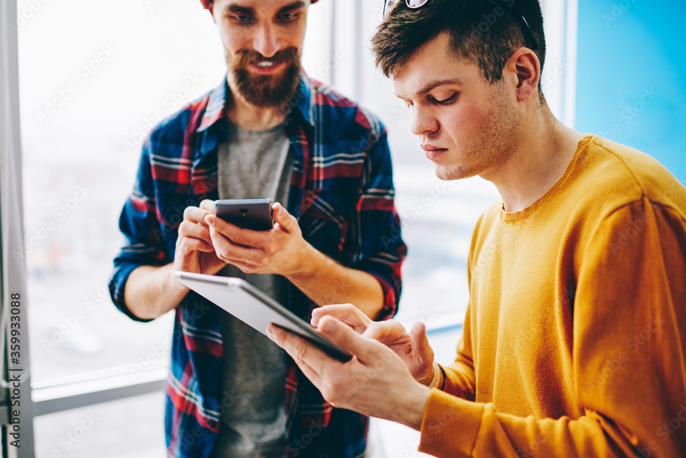 Poster pensive young man with modern touch pad in hand choosing media and sharing to positive bearded colle