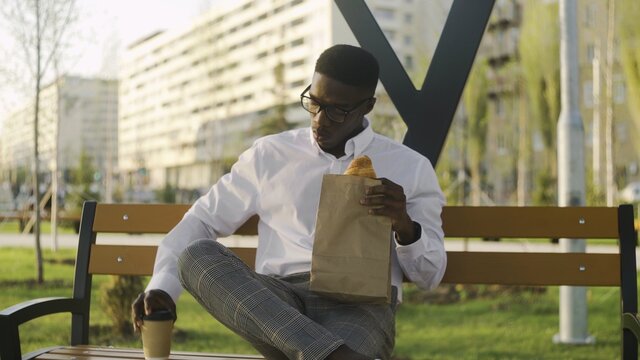 African american businessman relaxing and eating croissant with coffee on the bench in city park