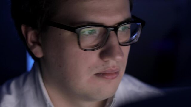 Man in eyeglasses late at night scrolling in front of laptop pan shot. Coder, programmer or developer using laptop in dark. Close up of glasses with reflection of computer screen.