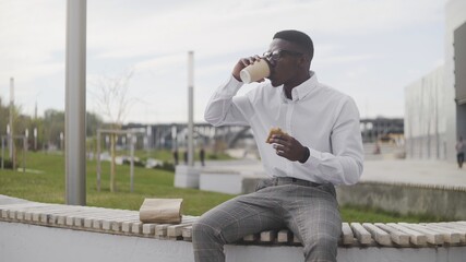 Fototapeta premium Young African American Man drinking coffee or tea and eating sandwitch in park near the office