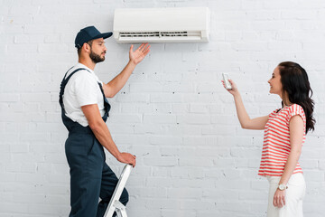 Side view of workman pointing at air conditioner near smiling woman holding remote controller - obrazy, fototapety, plakaty