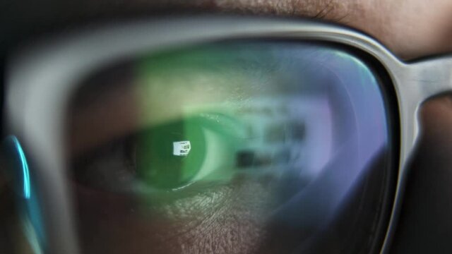 Male eyes in eyeglasses late at night scrolling in front of laptop. Coder, programmer or developer using laptop in dark. Close up of glasses with reflection of computer screen.