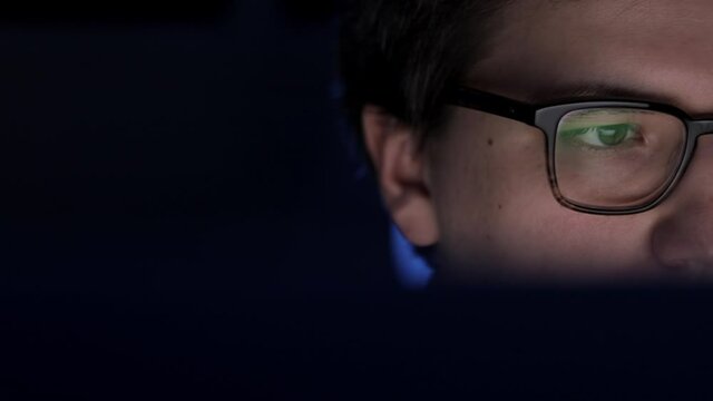 Man in eyeglasses late at night scrolling in front of laptop pan shot left to right. Coder, programmer or developer using laptop in dark. Close up of glasses with reflection of computer screen.