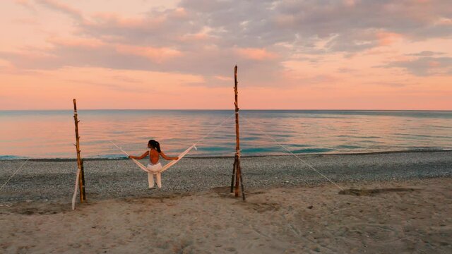 Bellissima ragazza va in vacanza sull'altalena in riva al mare calmo con nuvole e cielo arancione e rosso.