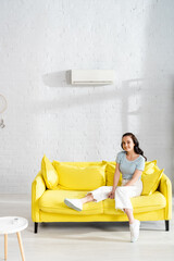 Young woman smiling at camera while sitting on sofa near remote controller of air conditioner on coffee table