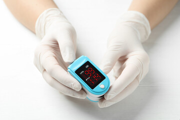 Doctor in gloves holding fingertip pulse oximeter at white wooden table, closeup