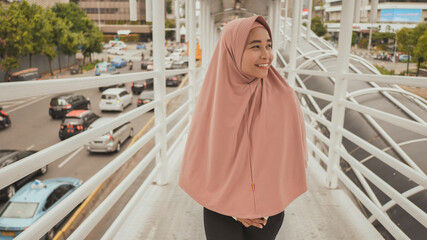 Smiling girl in hijab goes behind the camera on the transition over road traffic. Jakarta. Indonesia.