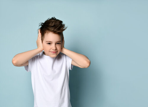 Sly naughty kid smiling and covering ears with hands showing he does not want to hear and obey. Studio portrait isolated on light blue
