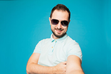 A young bearded man is fooling around at the camera on a blue background. The guy takes a selfie and builds funny faces