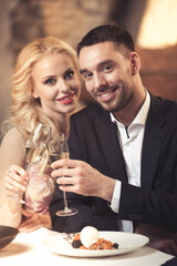 Romance and dating concept. Young couple man and woman with champagne glasses celebrating and toasting in restaurant. Two people male and female in elegant evening suit and dress on romantic dinner