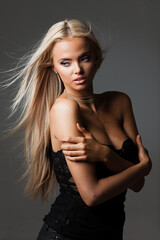 Fototapeta premium Portrait of blond young woman with beautiful makeup and hairstyle in a black lace dress stands on gray background. Fashion model poses in elegant clothes in studio