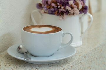 coffee with heart shaped milk froth near window with a vase with dried flower.