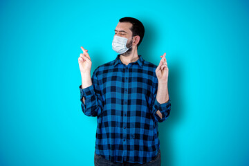 Closeup portrait young bearded man using mask and praying hands clasped hoping for best asking for forgiveness or miracle isolated blue wall background. Coronavirus concept