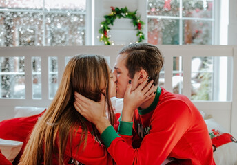 Young man and woman in red Christmas pyjamas kissing in a white room with big window. Snow background.