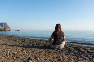 Fototapeta na wymiar The girl is sitting on the beach. Beautiful brunette girl alone against the background of a blue calm sea. Peace and contemplation. Enjoy the beautiful view. The concept of loneliness. Authenticity.