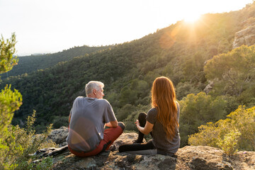 Back view of father and adult daughter sitting on rock in the mountains enjoying sunset