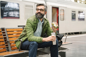 Man sitting on a bench at the train station with sandwich and hot drink