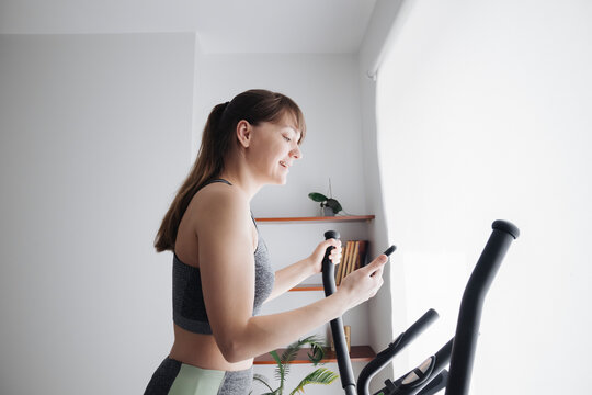Woman using smartphone while performing workout on elliptical trainer at home