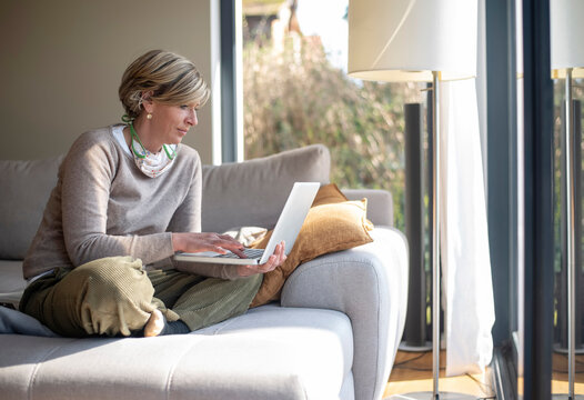 Woman Using Laptop While Sitting On Sofa In Living Room During Working From Home