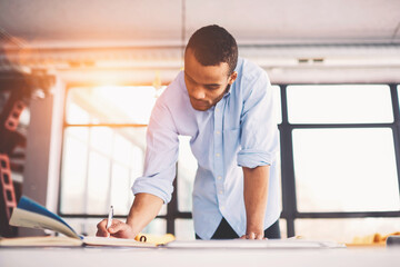 Skilled male afro american engineer thinking over blueprint improvement noting ideas in copy book to correct mistakes in planning looking concentrated on workspace desk with papers in modern studio