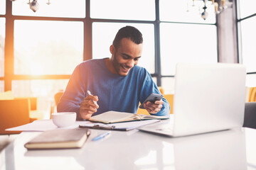Cheerful attractive afro american male freelancer chatting with friends in social networks via smartphone interrupting important job with updating software on laptop computer connected to wifi