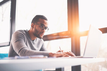 Concentrated dark skinned copywriter in eyewear typing text of publication for web site working on...