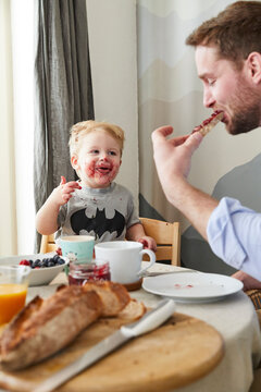 Portrait of smeared little boy at breakfast table watching his father eating bread with jam