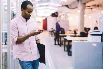 Young male employee waiting for meeting with colleagues while surfing internet and making online shopping in web store choosing digital devices via smartphone connected to fast 5G wireless in office