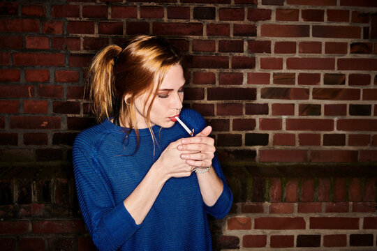 Young woman lighting cigarette against brick wall at night