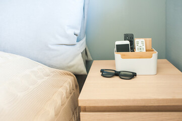 Organizer box for storing personal belongings, stands on the bedside table near the bed. Inside the phone, a remote control for light, near glasses.