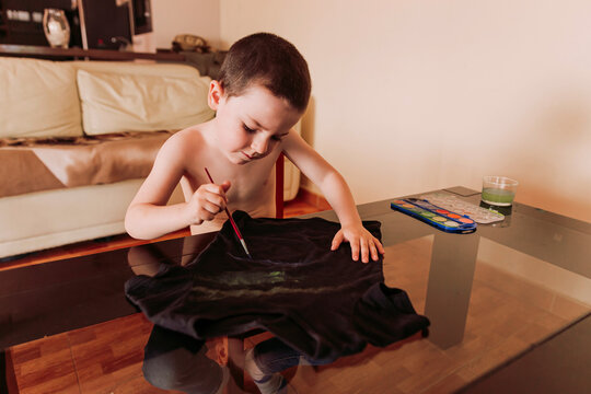 Shirtless boy drawing on black t-shirt at table in living room