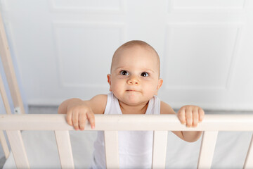 portrait of a smiling baby boy 8 months old standing in a crib in a children's room in white clothes and looking at the camera, morning baby, baby products concept