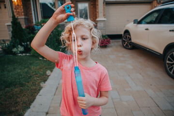 Young Caucasian girl blowing soap bubbles on home front yard. Child having fun outdoors on sunset....