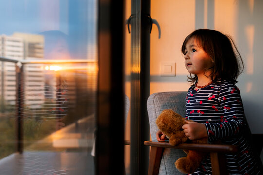 Portrait of little girl with teddy bear looking out of window at sunset