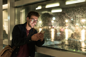 Smiling young man using smartphone at rainy window at night