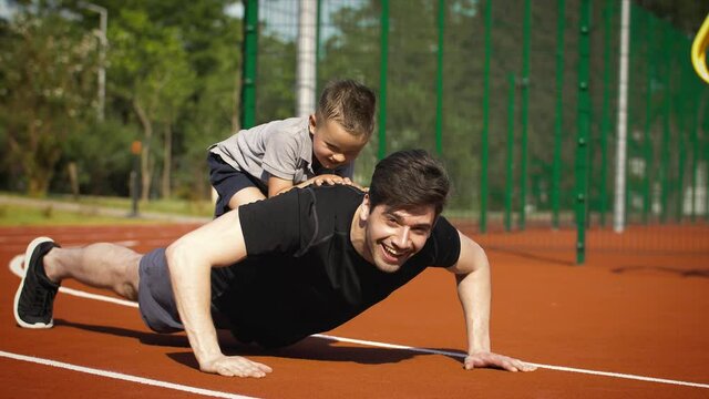 Dad in sportswear is doing push-ups while his little child boy is sitting on his back and laughing. Father spends time with his kid at sports ground